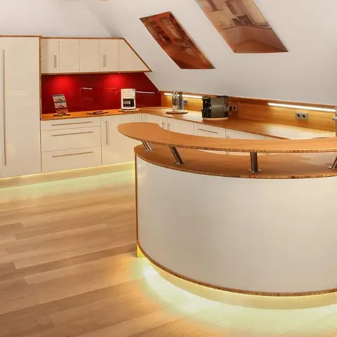 Attic kitchen under a sloped ceiling with glossy white cabinetry, a red glass splashback, and a curved wood-topped breakfast bar with underlit base.
