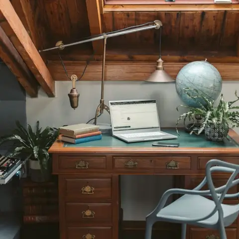 Attic study nook with a vintage wood desk under a sloped wood-beamed ceiling, a brass desk lamp, a globe and potted plants