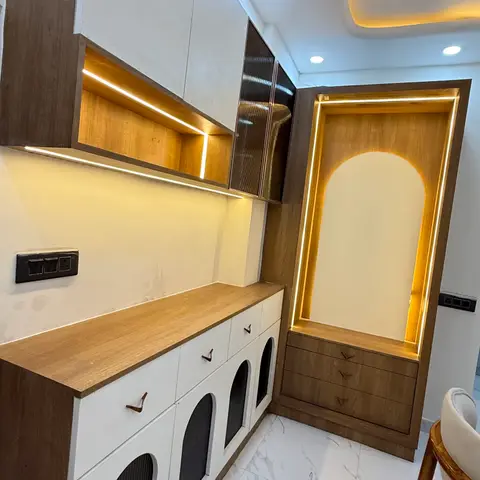 Backlit arched pooja niche framed in warm wood with profile LED lighting, set beside a white-and-wood crockery unit with arched fluted-glass drawer fronts