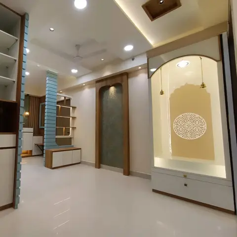 Backlit arched pooja unit with a carved Om medallion jaali panel and hanging brass bells, set beside a textured stone niche in an open hallway.