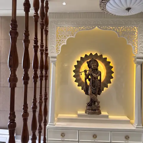 Backlit carved-arch pooja niche with a bronze flute-playing Krishna statue and matching drawer storage below, seen past turned wooden staircase spindles.