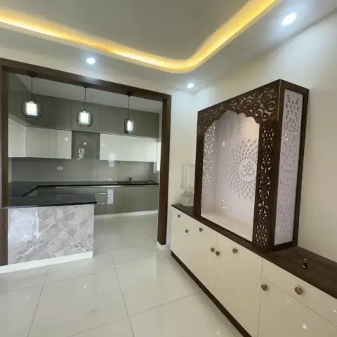 Backlit carved-wood jaali pooja unit with an Om motif beside a foyer opening onto a glossy modular kitchen with pendant lighting.