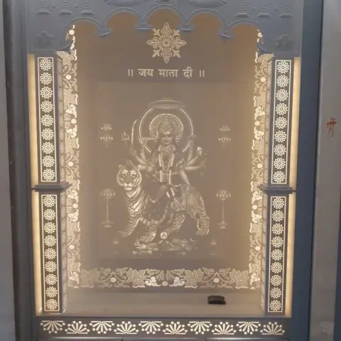 Backlit CNC-carved arched pooja mandir cabinet with an engraved Durga motif and Sanskrit inscription, fluted jaali side pillars lit from within.