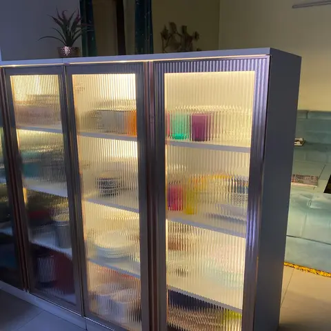 Backlit crockery display cabinet with fluted glass doors and colorful glassware, positioned in front of a mint velvet sofa set in the living room beyond.