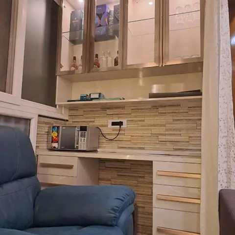Backlit glass-front bar cabinet with a stone-clad backsplash above a countertop with brass-handled drawers, beside a blue reclining armchair.