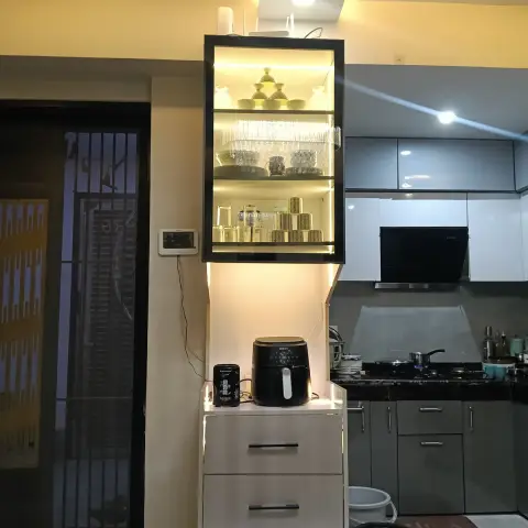 Backlit glass-front crockery display unit with a three-drawer base beside a grey modular kitchen counter and gas hob.