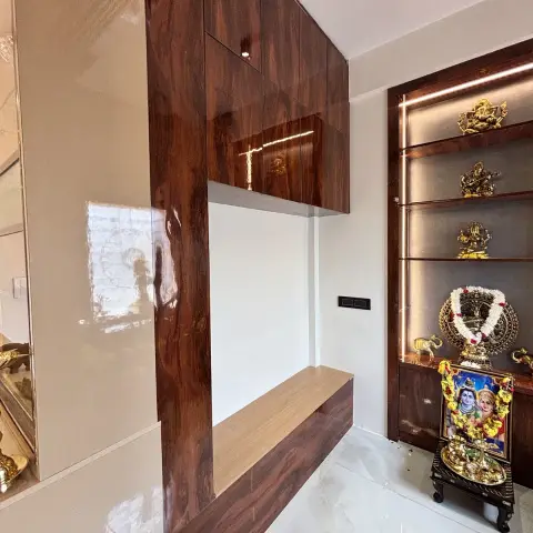 Backlit glass-shelf pooja unit in a dark wood-veneer frame displaying brass deity idols and a garlanded photo, beside a glossy wood-veneer wall and bench.