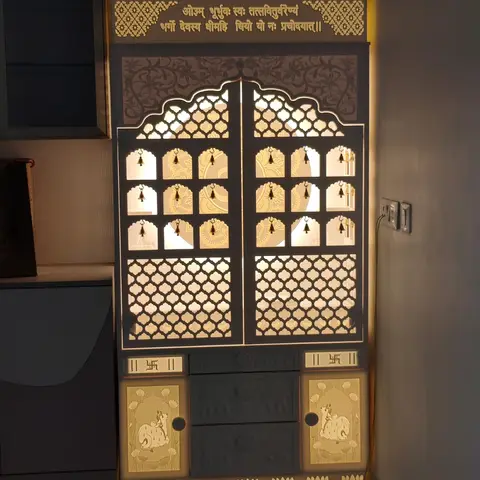 Backlit pooja cabinet with arched jaali-cut doors, brass bells, a Gayatri mantra inscription across the top and cow-motif panels on the lower doors.