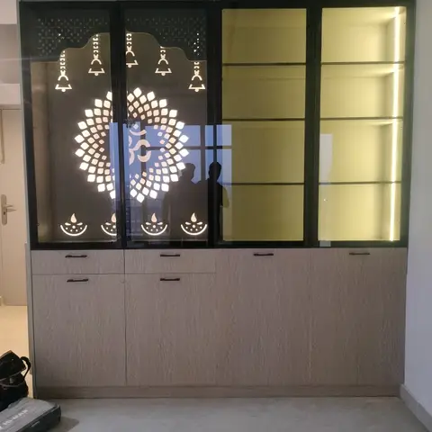 Backlit pooja unit with a laser-cut CNC jaali panel featuring an Om motif, hanging bells and diyas, over light wood-grain laminate storage cabinets.