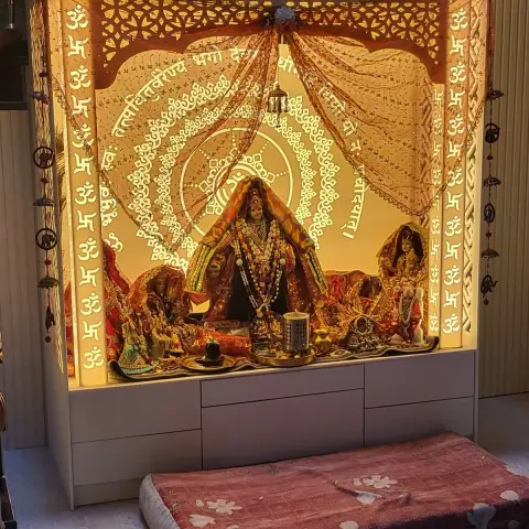 Backlit temple-style pooja unit with carved Om and Sanskrit motifs, sheer curtain drape, and deity idols, set beside a staircase.