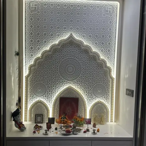 Backlit white CNC lattice pooja niche with a cusped Mughal arch and swastik corner medallions