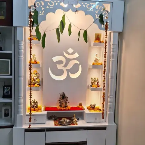 Backlit white pooja unit with a carved arched jaali crown around a glowing Om motif, turned pillars and marigold torans on either side.