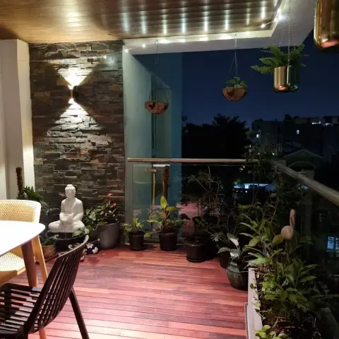 Balcony deck at night: a stone-clad wall lit by an up-down sconce behind a white Buddha water feature, potted foliage along a glass railing