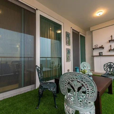 Balcony fitted with artificial turf and verdigris cast-iron outdoor table and chairs, with a brass idol display shelf on the back wall.
