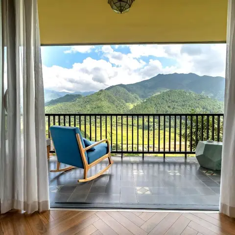 Balcony framed by sheer curtains with a teal wood-frame rocking chair overlooking a green mountain valley
