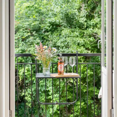Balcony nook with a fold-down black metal table set with a wine bottle, glasses and a wildflower arrangement, framed by open French doors and greenery.