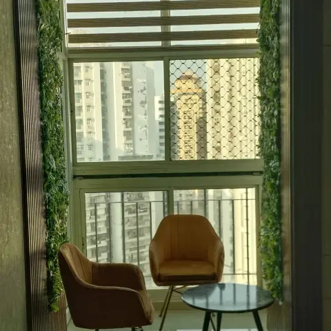 Balcony sit-out with two tan velvet accent chairs and a round marble-top table, framed by faux greenery and a high-rise city view.