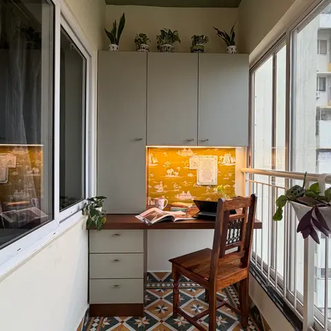 Balcony-turned-study nook with a wood desk against mustard elephant-motif wallpaper, a fluted grey wardrobe, patterned floor tiles, and a rattan pendant light.