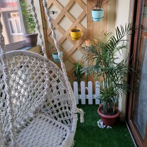Balcony with a macrame hanging swing chair, artificial turf underfoot, and a wooden lattice trellis holding colourful hanging planters.
