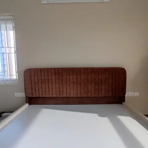 Bare beige bedroom wall with only a channel-tufted rust-brown upholstered headboard installed, mattress and window visible, no other furnishing yet.