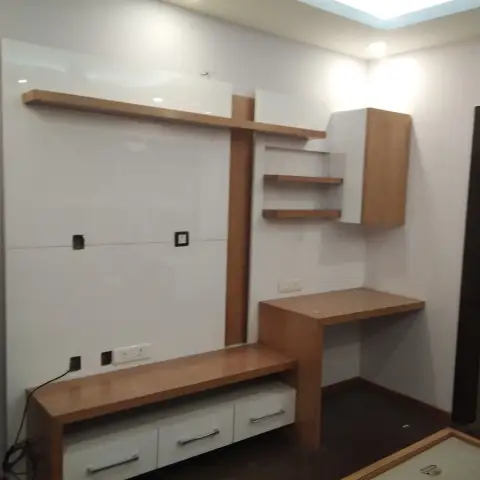 Bare study nook with a glossy white TV backdrop panel, wood floating shelves, and a built-in study desk beside a wall-mounted cabinet.