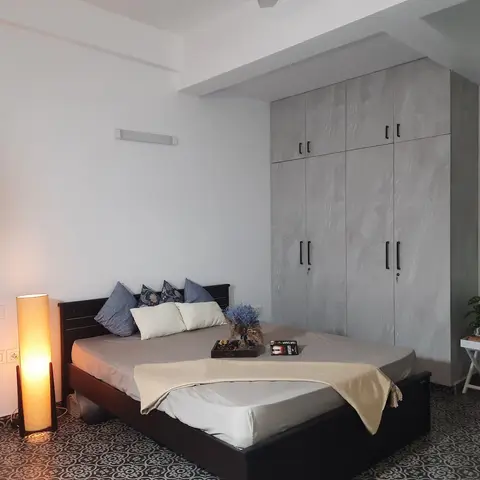 Bare white-walled bedroom over black-and-white patterned cement tile, with a floor-to-ceiling grey stone-look laminate wardrobe and a dark wood bed still