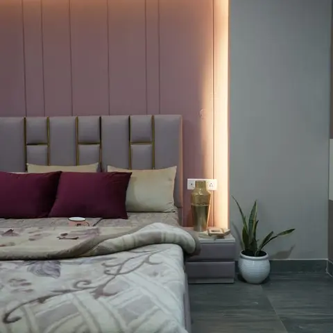 Bed corner with a grey leatherette headboard divided by brass inlay strips, set against a dusty-pink panelled recess washed by a concealed warm strip