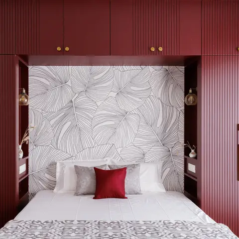 Bed nook framed by a fluted maroon wraparound wardrobe wall with brass handles, set against a monstera-leaf line-art wallpaper.