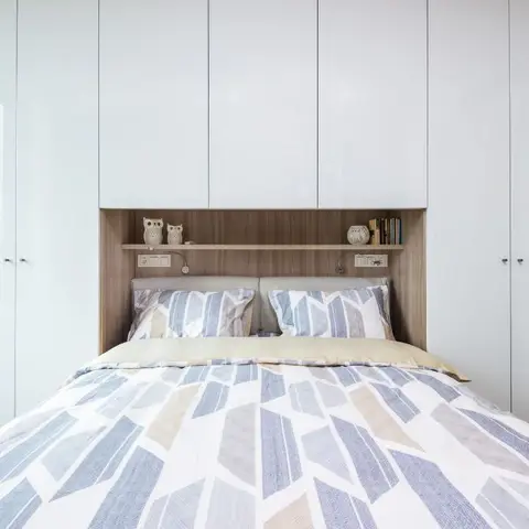 Bed slotted into a floor-to-ceiling white gloss wardrobe wall with an oak-lined overhead bridge shelf holding two owl figurines and a row of books