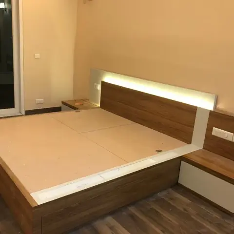 Bed under construction showing a wood-veneer platform frame with an integrated LED-backlit headboard and a floating attached nightstand.