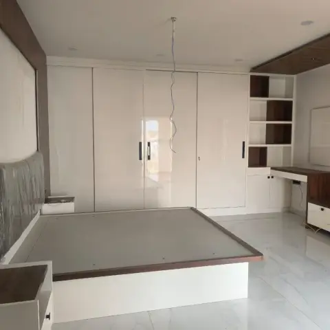 Bedroom at handover: white platform bed with a walnut-lipped base and its headboard still in plastic wrap, full-height gloss sliding wardrobe