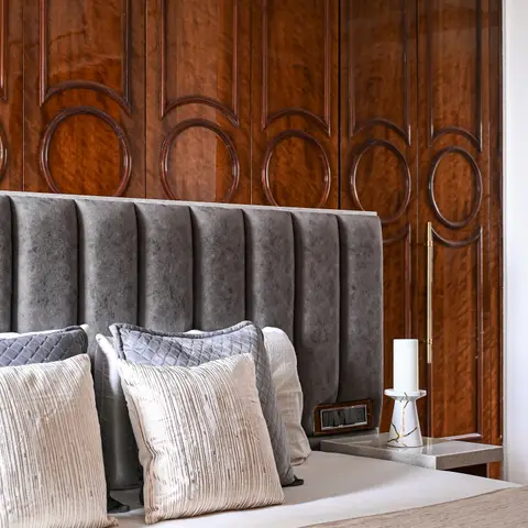 Bedroom close-up of a channel-tufted grey headboard with layered cushions against a dark wood wardrobe featuring carved circular panel mouldings