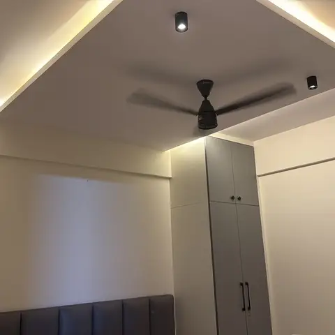 Bedroom corner showing a stepped, cove-lit POP ceiling with a ceiling fan and a grey-shuttered wardrobe beside an upholstered headboard.