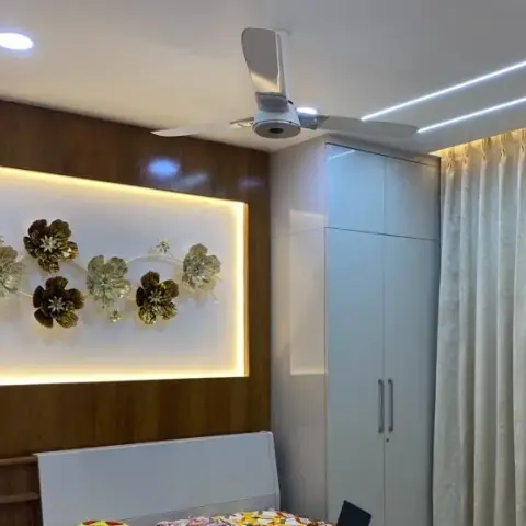Bedroom corner where a backlit recessed panel set into wood cladding holds a spray of gold and brown metal flowers, with a white gloss wardrobe alongside