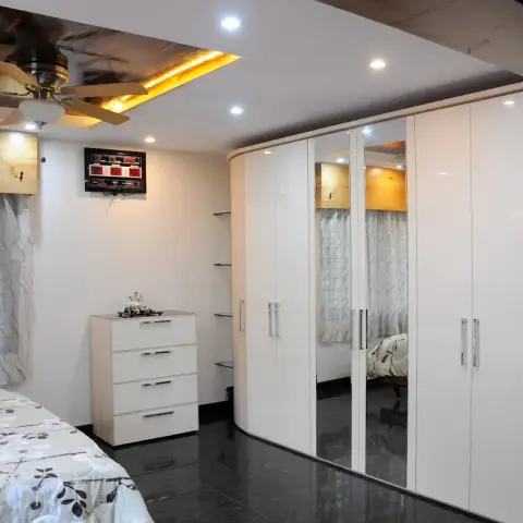 Bedroom corner where a curved-end white gloss wardrobe with two mirrored shutters wraps the wall beside a four-drawer chest