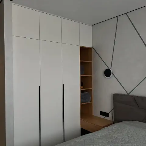 Bedroom corner where a matt white floor-to-ceiling wardrobe with recessed black handles meets a grey micro-cement wall scored with thin metal inlay lines