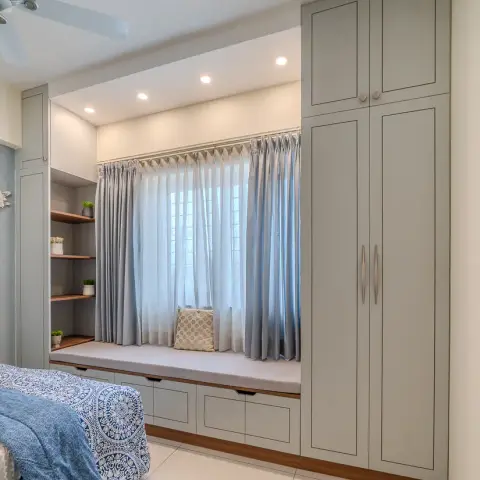 Bedroom corner where a sage-grey shaker wardrobe with loft turns into a cushioned window seat over four drawer fronts