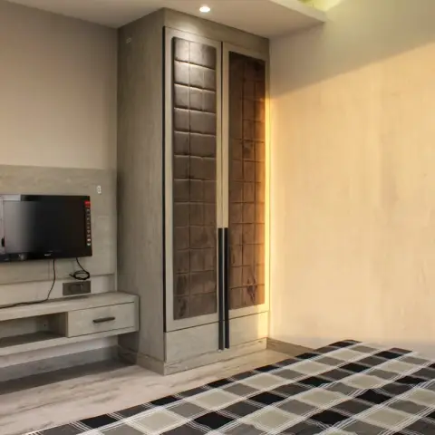 Bedroom corner where a small TV hangs on a pale-wood laminate backing panel above a floating drawer console
