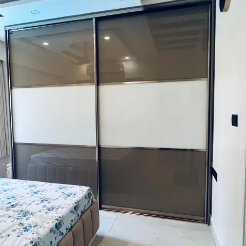 Bedroom corner where a two-shutter sliding wardrobe in bronze-tinted and white lacquered glass stands a step from the upholstered bed