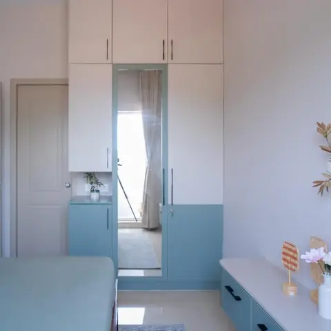 Bedroom corner with a sage-and-white wardrobe featuring a full-length mirror door, paired with a low console styled with dried florals and a vase.