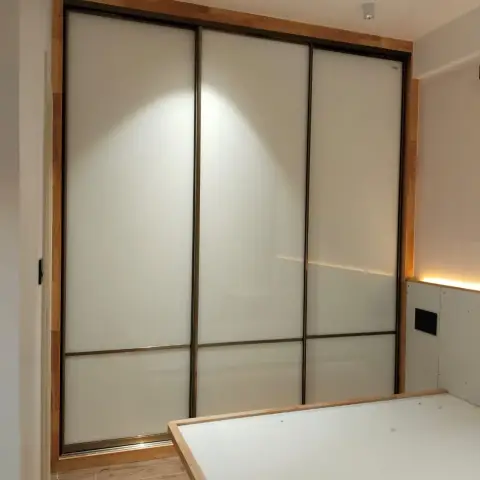 Bedroom in progress with a floor-to-ceiling frosted-glass sliding wardrobe framed in oak and a white bed platform base awaiting its headboard