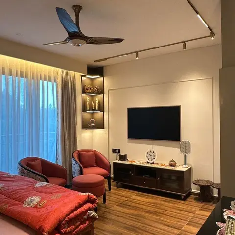 Bedroom lounge corner with rust-red barrel armchairs and a matching ottoman facing a wall-mounted TV over a dark wood console, under a linear brass track light.