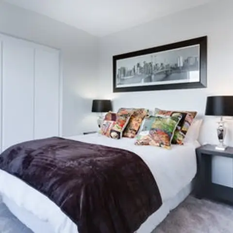Bedroom on grey carpet with a chocolate faux-fur throw folded across a white bed, patterned collage cushions