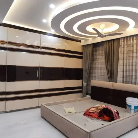 Bedroom photographed at handover, bare bed base still holding paint buckets and cloth, under concentric circular POP rings that ring the ceiling fan