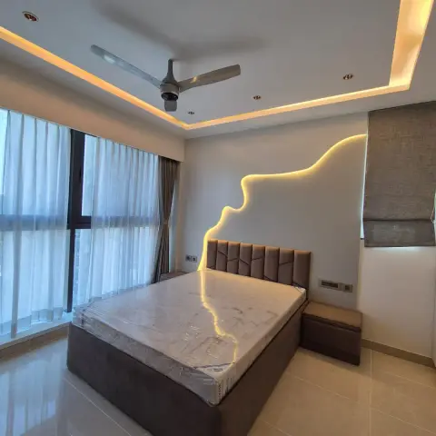 Bedroom photographed at handover with the mattress still shrink-wrapped: taupe upholstered storage bed and matching side stool under a wavy concealed-LED line