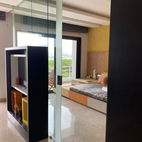 Bedroom seen from the passage through a frameless glass partition: a low platform bed with a grey velvet throw and orange bolsters against a jute-textured