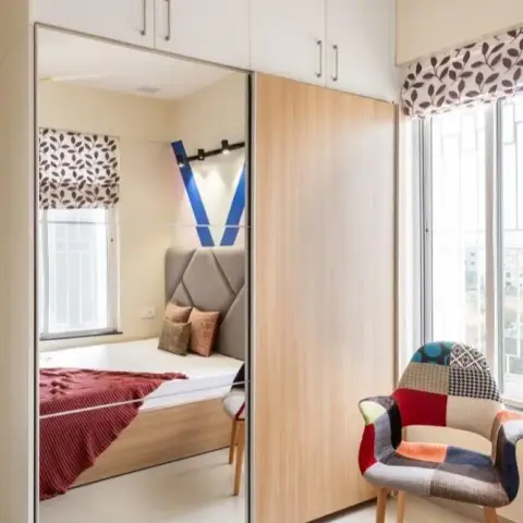 Bedroom shot along a two-track sliding wardrobe whose mirror leaf reflects the bed's grey faceted upholstered headboard and red throw