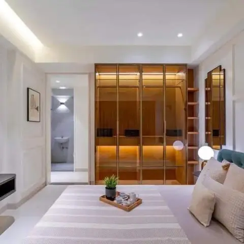 Bedroom shot from the foot of a teal button-tufted bed, looking at a bronze-glass walk-in wardrobe lit from inside