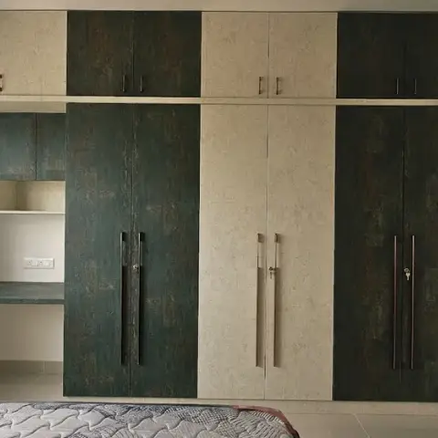 Bedroom shot low across the foot of a bare mattress at a full-wall wardrobe run alternating oxidised-teal and beige stone-textured laminate shutters with lofts