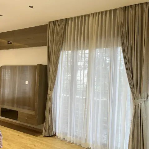 Bedroom shot square at the window wall, where full-height beige pinch-pleat drapes over white sheers meet a curved-top veneer TV panel on a floating console